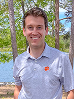A smiling man in a gray polo shirt with a Clemson University logo stands by a lake surrounded by trees.