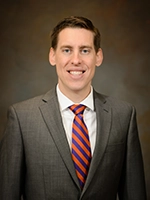 Studio portrait of a smiling man in a gray suit with a white shirt and red-and-blue striped tie against a brown backdrop.