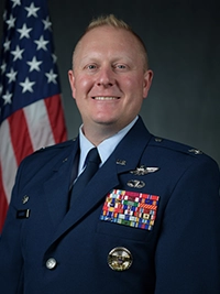 Formal portrait of a uniformed U.S. Air Force officer with medal ribbons, standing in front of an American flag backdrop.