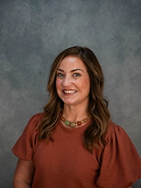 Smiling woman with long brown hair wearing a rust-colored blouse and beaded necklace, set against a textured gray background.
