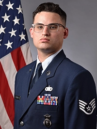 Formal portrait of a uniformed U.S. Air Force service member with medals and insignia, standing in front of an American flag.