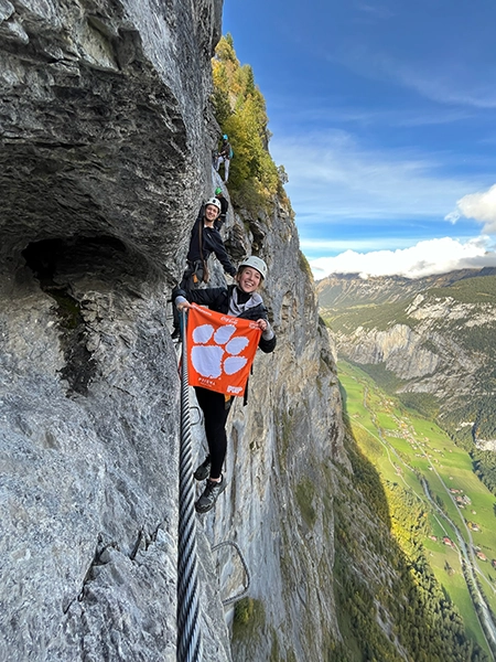 Two climbers on a narrow rock face proudly display a Clemson University flag against a backdrop of stunning mountain scenery.