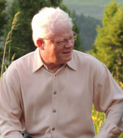 A smiling man with silver hair sitting outdoors in a light-colored button-up shirt amidst greenery.