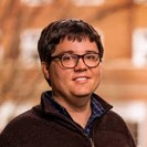 A portrait of a smiling individual with glasses, wearing a dark sweater and a plaid shirt. The background features an out-of-focus brick building.