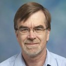 Professional headshot of a middle-aged man with glasses and gray hair, wearing a light blue shirt.