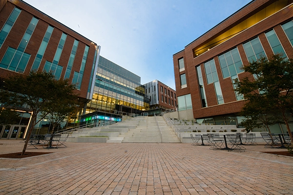 Modern university campus buildings with large glass windows and a central staircase. The image showcases an inviting outdoor plaza area with seating.