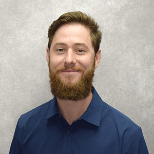 Professional portrait of a man with a beard, wearing a blue shirt and smiling against a neutral background.