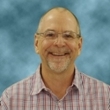 Headshot of a smiling man with glasses, wearing a plaid shirt against a blue background.
