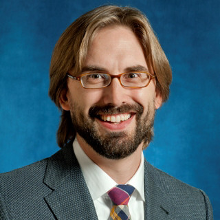 Headshot of a smiling man with glasses and a beard, wearing a suit and tie against a blue background.
