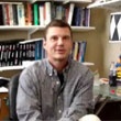 Portrait of a man seated in an office with shelves of books and binders in the background.