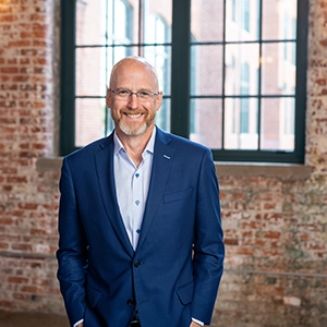 Smiling bald man with glasses in a blue suit standing indoors in front of large industrial-style windows and a brick wall.