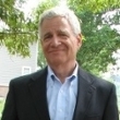 A smiling man in a suit with gray hair stands outdoors, framed by green trees.