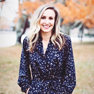 Smiling woman with long blonde hair wearing a navy floral dress, standing outdoors with autumn trees in the background.