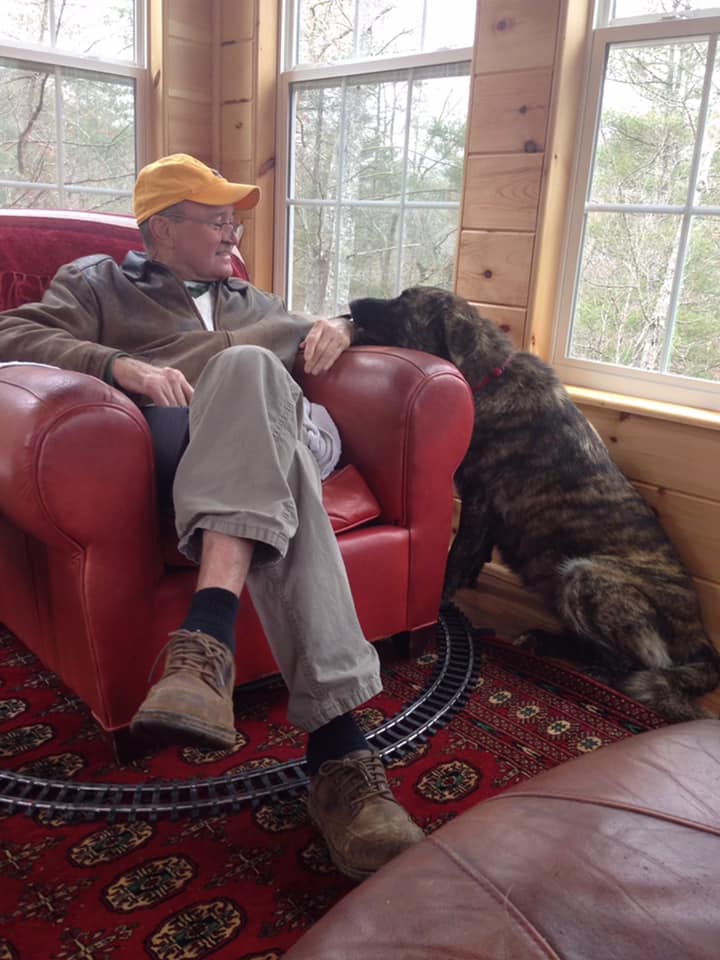 A man in a yellow cap relaxes in a red armchair while interacting affectionately with a large brindle dog beside him.