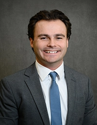 Headshot of a young man in a dark suit and white shirt, smiling against a gray backdrop.