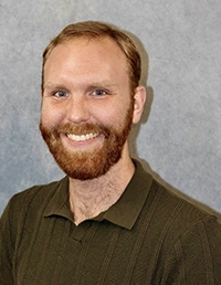 Smiling bearded man in a dark polo shirt against a gray studio backdrop.