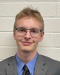 Portrait of a smiling young man wearing glasses, a gray suit jacket, blue shirt, and dark tie in front of a light brick wall.