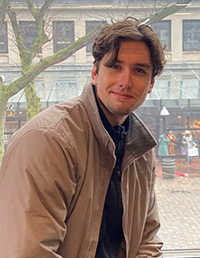 A man with wavy brown hair, wearing a beige jacket, smiles while sitting by a window with a view of a bustling street and shops.