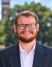 Smiling bearded man wearing glasses and a dark blazer, standing outdoors with trees in the background.