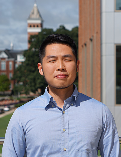 A young man wearing a blue dress shirt stands outdoors with a university building and clock tower visible in the background.