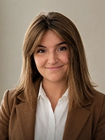 Smiling person with shoulder-length brown hair wearing a brown blazer over a white shirt, posed against a plain light background.
