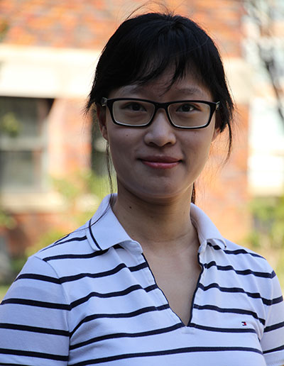 A young woman with glasses stands outdoors, wearing a striped polo shirt, smiling at the camera.