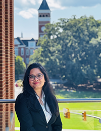 Woman in a black blazer and glasses standing outdoors on a balcony, with green lawn, trees, and a clock tower in the background.