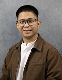 Studio headshot of a person wearing glasses, a brown jacket, and a white shirt against a gray background.
