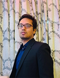 Person in a black blazer and glasses, standing indoors in front of patterned curtains and looking to the side.