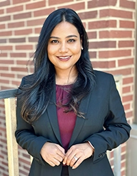 Smiling woman in a dark blazer over a maroon top, standing in front of a brick wall.