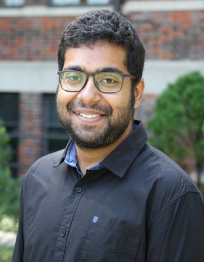 Smiling young man with glasses and a beard, wearing a black shirt, posed outdoors against a brick building background.