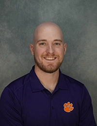 Professional headshot of a man with a bald head, smiling, wearing a purple polo shirt with a Clemson University logo.