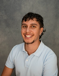 Smiling young man with curly hair wearing a light blue polo shirt against a gray background.