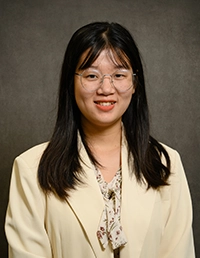 Professional headshot of a young woman with glasses, wearing a light-colored blazer and a patterned blouse.