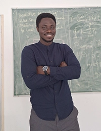 Smiling man standing with arms crossed in front of a chalkboard covered with writing.