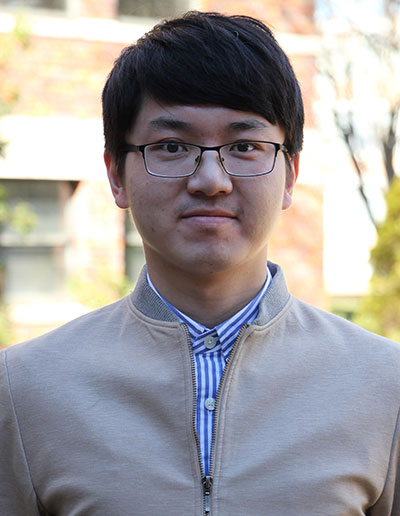 A young man wearing a beige jacket and glasses, standing outdoors with a subtle smile.