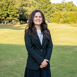 A woman in a black blazer smiling while standing on a grassy lawn.