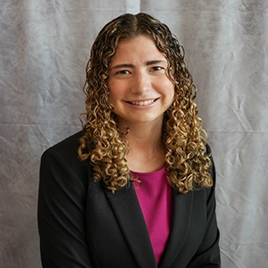 Professional portrait of a woman with curly hair wearing a blazer and smiling against a gray backdrop.