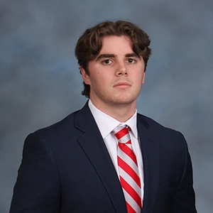 A formal portrait of a young man wearing a dark suit with a white shirt and red-striped tie, set against a soft blue background.