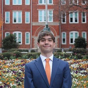 A young man in a suit with an orange tie stands confidently in front of a building, surrounded by colorful flowers.