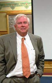 A man in a suit with an orange tie stands confidently in front of a blank presentation screen.