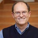 Headshot of a smiling man with glasses, wearing a blue shirt and dark vest, against a wooden staircase background.