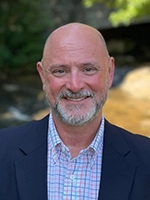 Head-and-shoulders portrait of a smiling bald man with a gray beard wearing a navy blazer and checkered shirt, with a blurred outdoor background.