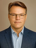 Professional headshot of a man with short, styled hair wearing glasses and a blazer.
