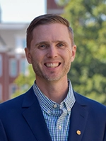 A professional headshot of a man in a navy blazer and checkered shirt, smiling against a blurred outdoor background.