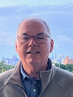 A smiling man wearing glasses and a light gray sweater, with a city skyline in the background.