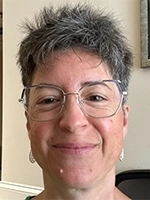 Close-up headshot of a smiling person with short spiky gray hair and large eyeglasses against a plain background.