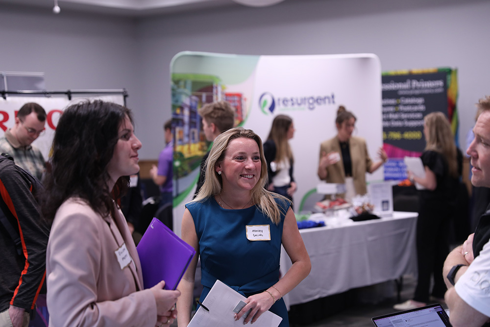A bustling trade show featuring various vendors with colorful displays, including tables with logos for Mimaki and MacDermid Graphics Solutions.
