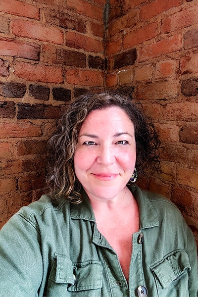 A smiling woman with curly hair, wearing a green jacket, stands in a cozy brick corner.