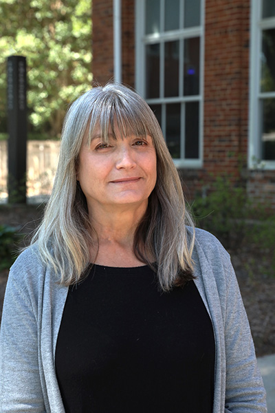 A woman with long gray hair stands outside a brick building, wearing a black top and gray cardigan.